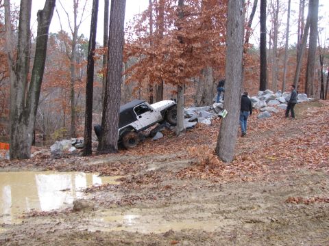 1979 Ford Bronco - A white Jeep Wrangler is shown attempting to traverse a challenging off-road course with muddy terrain and scattered rocks.