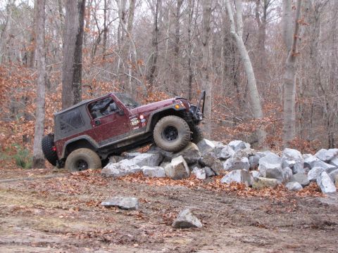 1979 Ford Bronco - A maroon Jeep Wrangler TJ navigates a rocky obstacle in a wooded area.