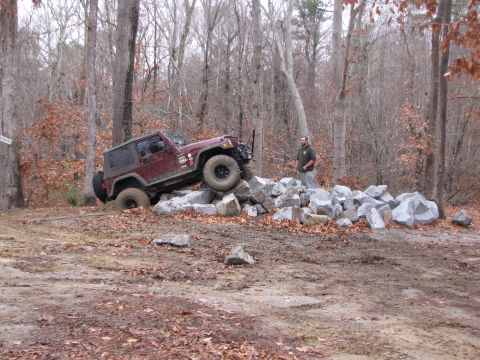 1979 Ford Bronco - A maroon Jeep Wrangler navigates a rocky obstacle course in a wooded area.