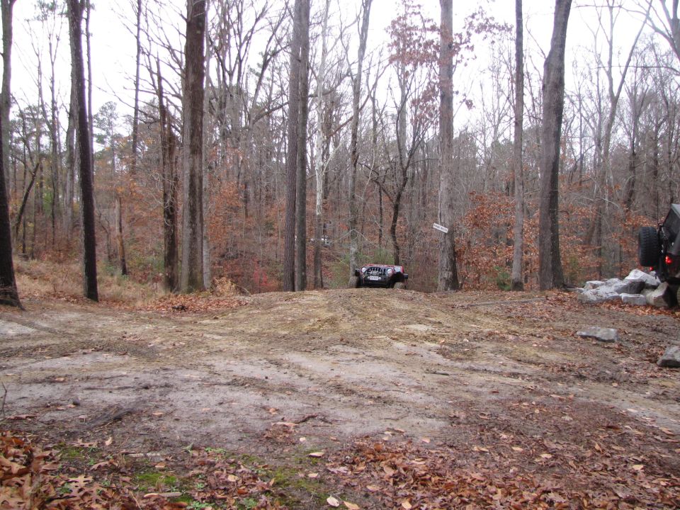 Jeep Wrangler on dirt hill, another vehicle visible on right. Woods in background