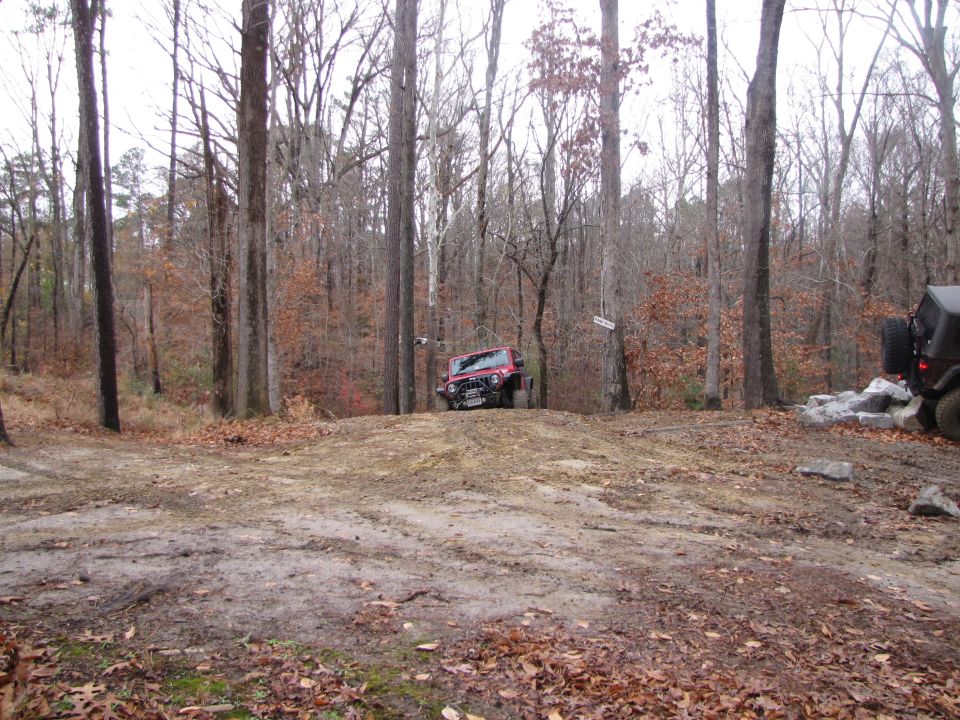 red Jeep Wrangler driving up a dirt hill in a wooded area, another Jeep is parked to the right