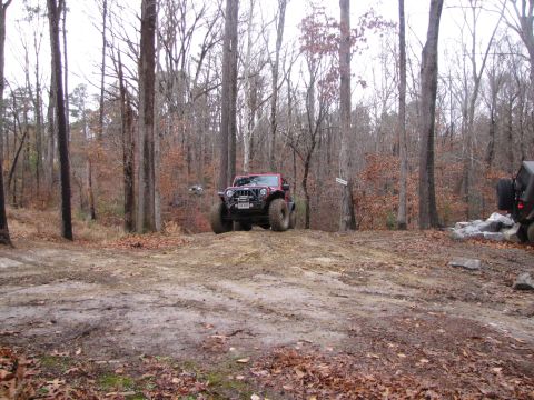 1979 Ford Bronco - A red Jeep Wrangler navigates an off-road trail in a forest, with a second vehicle partially visible.