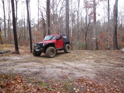 1979 Ford Bronco - A red Jeep Wrangler is parked on a muddy trail surrounded by trees.
