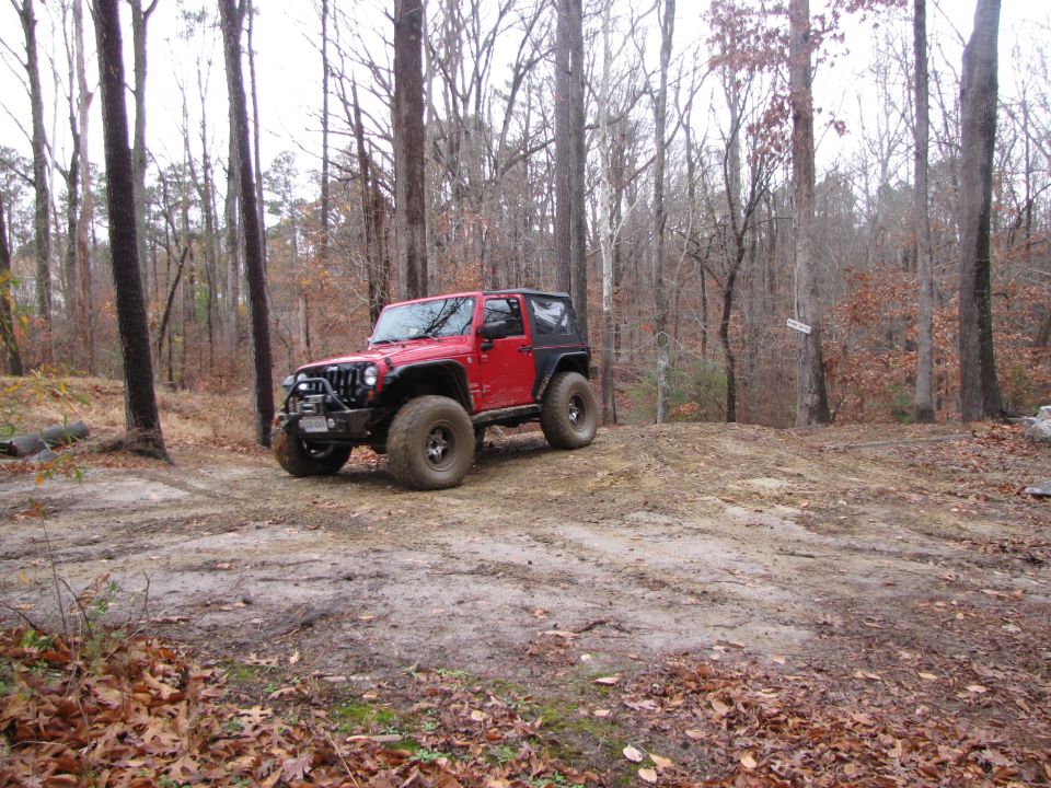 Red Jeep Wrangler with off-road tires on a dirt path in a wooded area