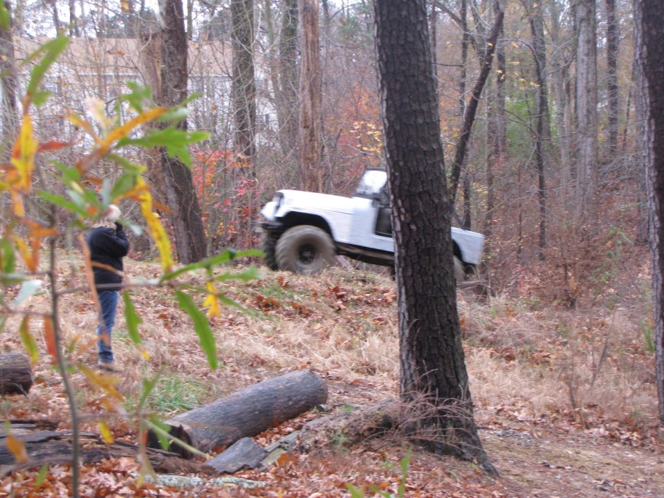 White off-road vehicle with large tires climbing a wooded hill. Person stands to the left