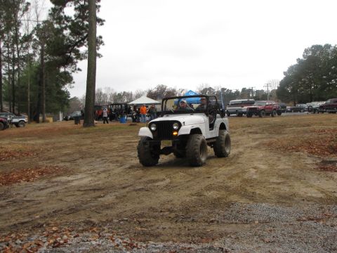1979 Ford Bronco - A white Jeep CJ with large off-road tires navigates a muddy track, with other vehicles and people gathered in the background.