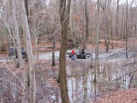 1979 Ford Bronco - Off-road vehicles traverse a wet and muddy path through a dense forest.