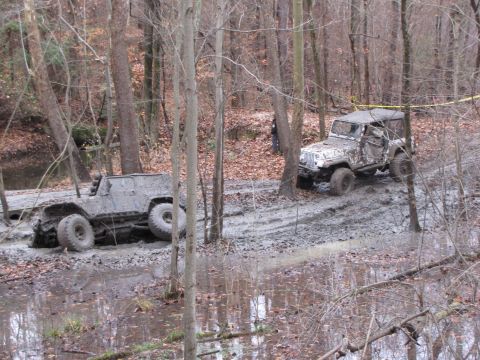 1979 Ford Bronco - Two off-road vehicles navigate a challenging muddy trail in a wooded environment.