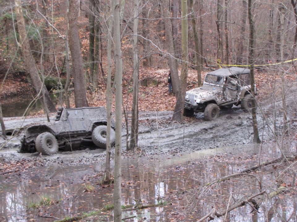 Two modified off-road vehicles covered in mud, one partially submerged in a muddy ditch, the other on a muddy trail in a wooded area