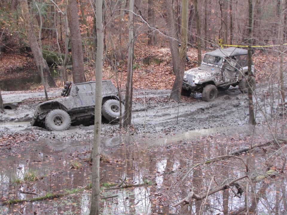 Two muddy off-road vehicles navigating a wooded trail with deep mud and water
