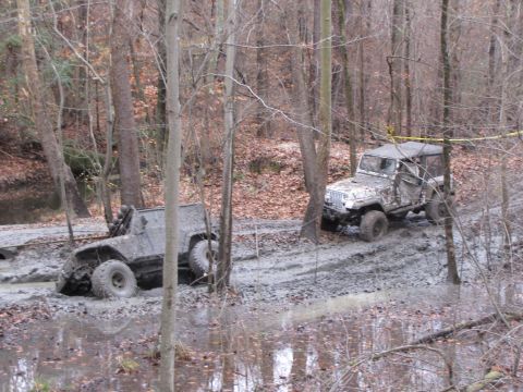 1979 Ford Bronco - Two off-road vehicles are stuck in deep mud on a wooded trail during an overcast day.