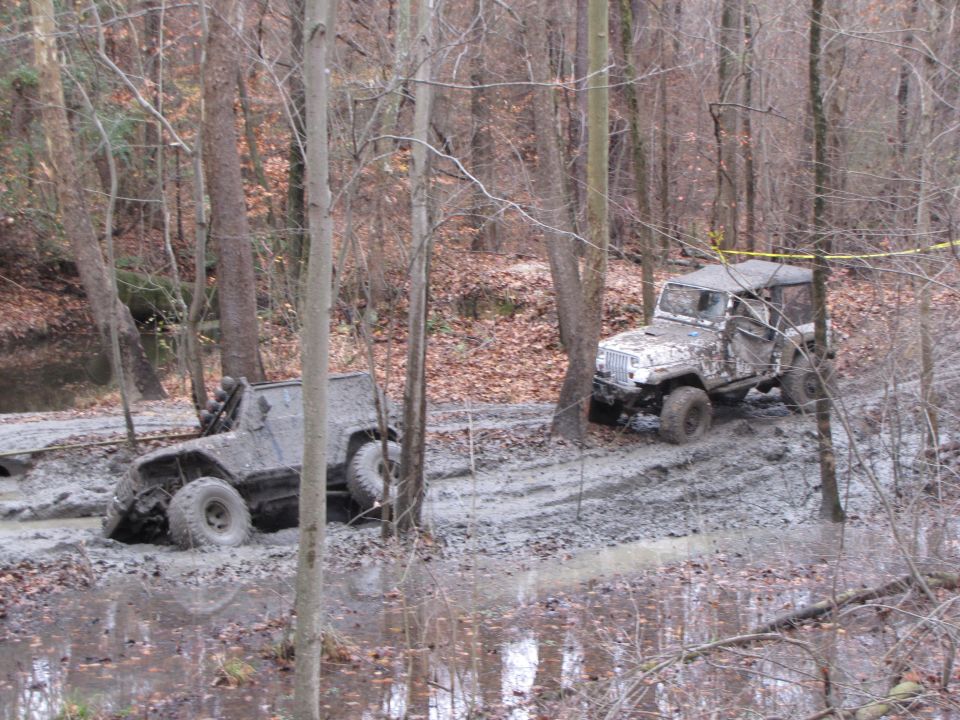 Mud-covered Jeep Wrangler and another off-road vehicle partially submerged in a muddy forest trail