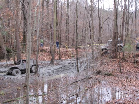 1979 Ford Bronco - Two off-road vehicles are stuck in deep mud and water in a wooded area, with people observing.
