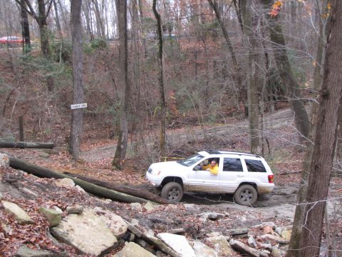 1979 Ford Bronco - White Jeep Grand Cherokee tackles a challenging off-road course in a wooded area.