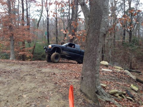 1979 Ford Bronco - A black pickup truck navigates an off-road trail through a wooded area with fallen leaves on the ground.