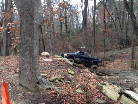 1979 Ford Bronco - A black pickup truck tackles a challenging off-road course through a wooded area with fallen leaves.