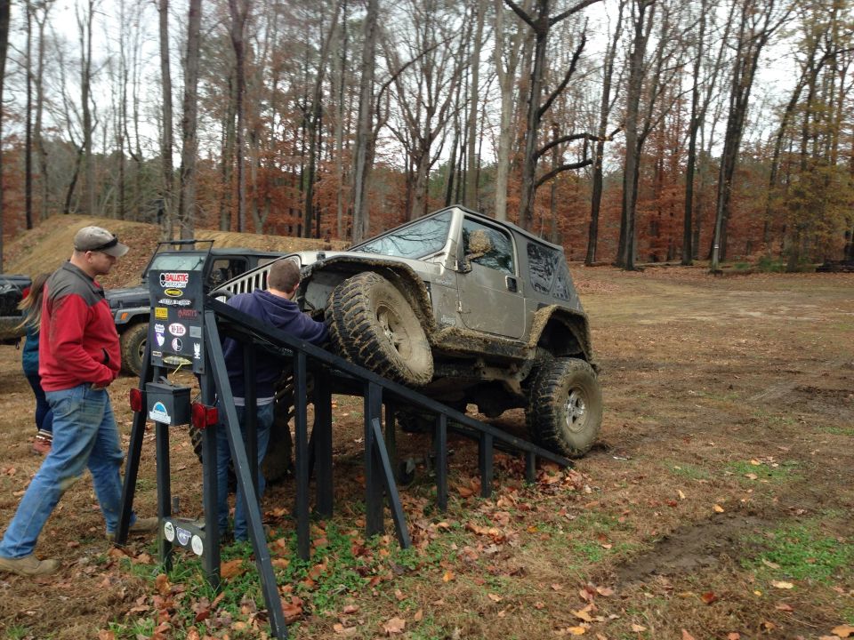 Silver Jeep Wrangler on a metal ramp, with two men nearby. Muddy terrain and trees in background