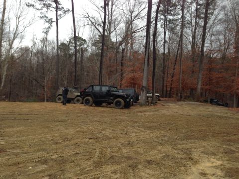 1979 Ford Bronco - A collection of off-road vehicles are parked in a clearing within a forest, with bare trees indicating a cooler season.