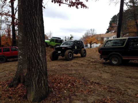 1979 Ford Bronco - Several off-road vehicles are parked on a muddy hill with trees and houses in the background.