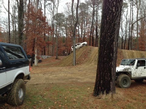 1979 Ford Bronco - A black and white SUV is parked in a wooded area, while a white Jeep and another vehicle navigate a dirt ramp in the background.