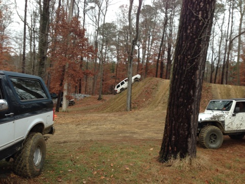 1979 Ford Bronco - Off-road vehicles navigate a dirt track in a wooded area. The black and white SUV is in the foreground.