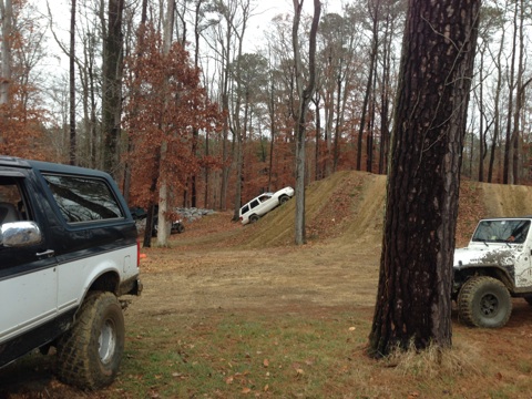 1979 Ford Bronco - Multiple off-road vehicles are seen on a dirt track in a wooded area, with one SUV attempting to climb a steep incline.