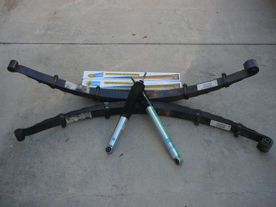 Two leaf springs and two Bilstein shock absorbers laid out on concrete. Boxes for the shocks are visible