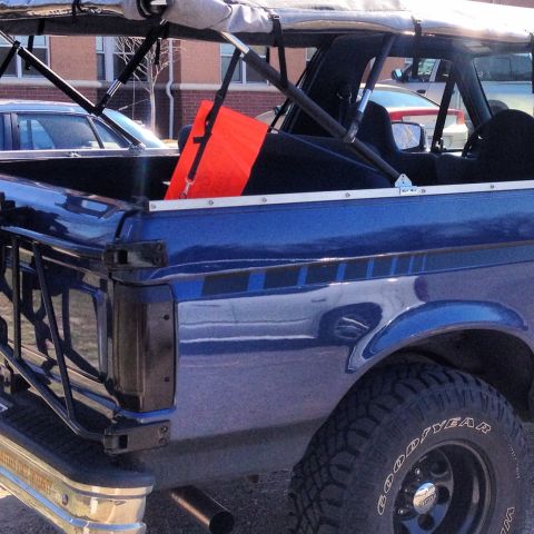 1996 Ford Bronco - The rear driver's side of a dark blue 1996 Ford Bronco is shown, with a bright orange safety flag visible in the bed. The vehicle has a rol…