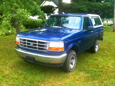 1996 Ford Bronco - A blue 1996 Ford Bronco is parked on a grassy lawn in front of a house and trees. The vehicle is viewed from the front three-quarter angle.