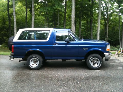 1996 Ford Bronco - A blue 1996 Ford Bronco with a white hardtop is parked on a paved surface in front of a wooded area. The side profile of the SUV shows the…