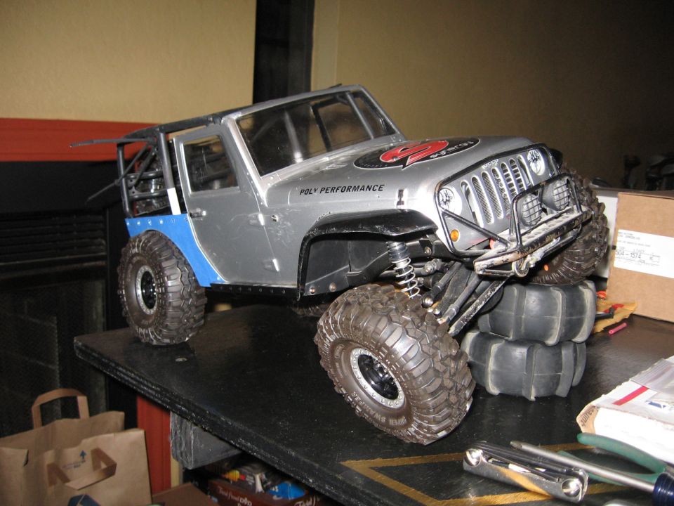 Silver RC rock crawler Jeep Wrangler with blue fenders and large tires.