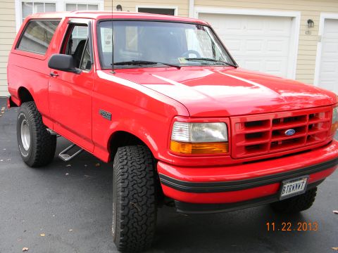 1995 Ford Bronco - A bright red 1995 Ford Bronco is parked on a paved driveway in front of a garage. The front quarter view shows the vehicle's grille, headli…