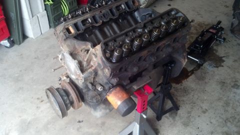 1988 Ford Bronco - A V8 engine block is supported by jack stands on a concrete floor. A black hydraulic jack is positioned near the engine, and there are pudd…