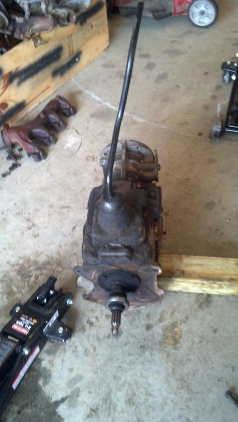 1988 Ford Bronco - A dirty manual transmission is shown on a concrete floor, possibly in a garage or workshop. A black gear shifter lever extends upwards from…