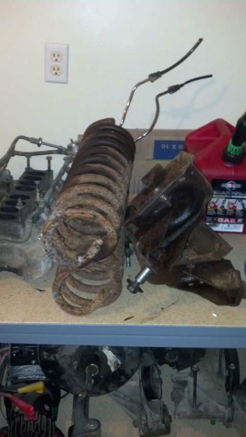 1988 Ford Bronco - Two heavily rusted coil springs and a dark metal suspension component sit on a workbench. An engine block and a red gas can are visible in…