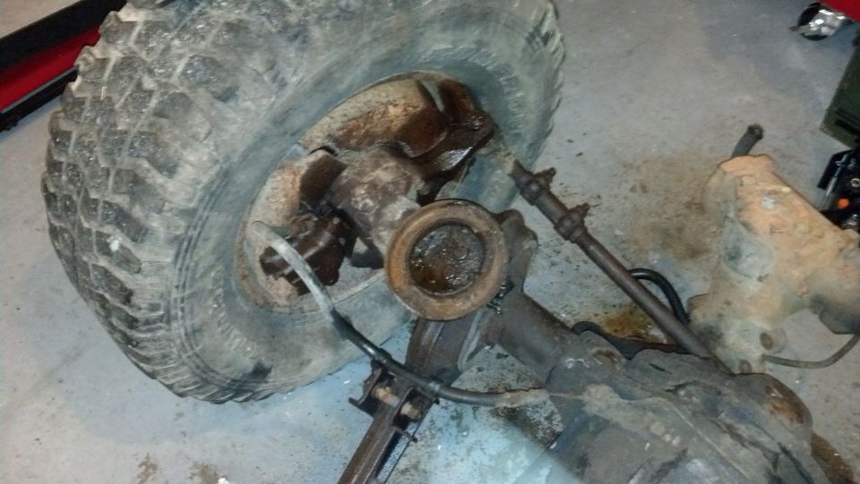 Front axle assembly with a large, worn tire, showing rust and grease. Visible steering linkage