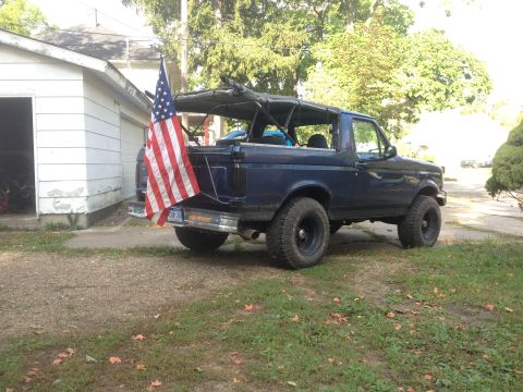 1996 Ford Bronco - This dark blue SUV, likely a 1992 model, is parked in front of a garage with an American flag flying from its rear.