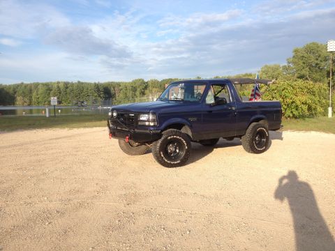 1996 Ford Bronco - A blue Ford Bronco is parked on a gravel lot with a lake and trees in the background.