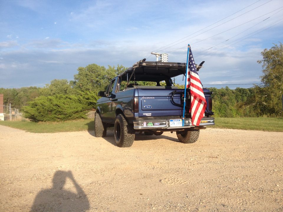 Blue Ford Bronco with a roof rack and an American flag attached to the rear bumper, seen from a rear 3/4 angle
