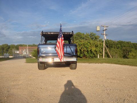 1996 Ford Bronco - Rear view of a blue SUV flying an American flag, parked on a gravel surface with trees and power lines in the background.