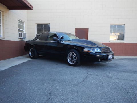 2003 Ford Crown Victoria - black full-size sedan with custom mesh grille and gray wheels parked at an angle beside a tan commercial building.
