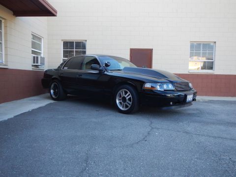2003 Ford Crown Victoria - Front-right view of a black full-size sedan with a chrome billet grille and gray five-spoke wheels in a parking lot.