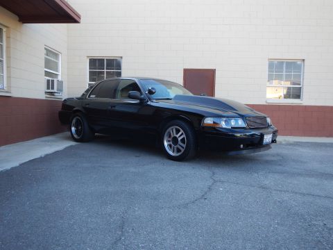 2003 Ford Crown Victoria - black full-size sedan with custom grille and tinted windows parked at an angle near a tan building.