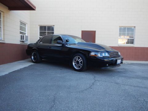 2003 Ford Crown Victoria - Three-quarter front view of a black full-size sedan with mesh grille and gray wheels beside a cream wall.