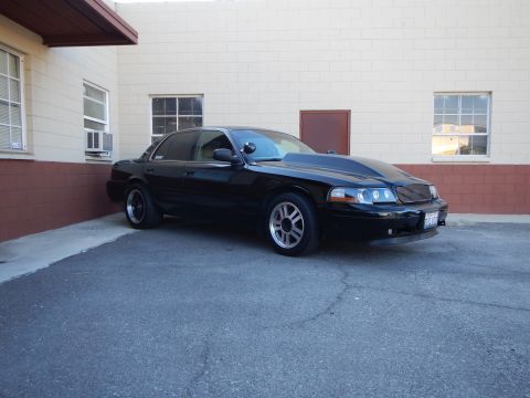 2003 Ford Crown Victoria - black full-size sedan with halo-ring headlights and a custom grille parked beside a tan commercial building.