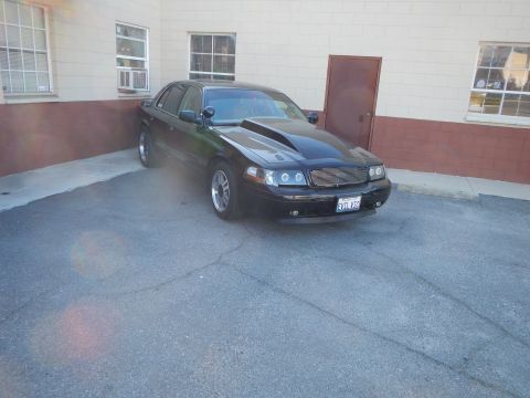 2003 Ford Crown Victoria - Front three-quarter view of a black full-size sedan with custom grille and California plate, with sun lens flare.