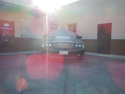 2003 Ford Crown Victoria - Head-on view of a black sedan backlit by heavy sun glare, showing a California plate reading EVIL VIC.