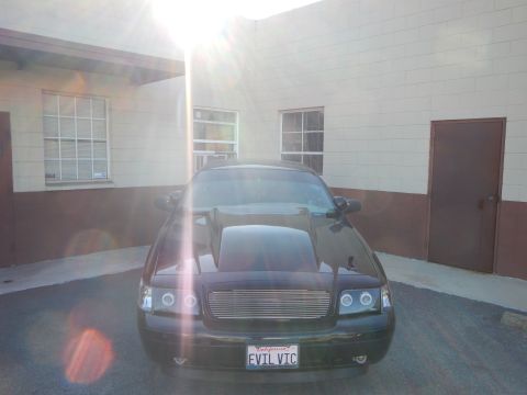 2003 Ford Crown Victoria - black full-size sedan parked head-on in a lot with intense sun glare above, EVIL VIC plate visible.