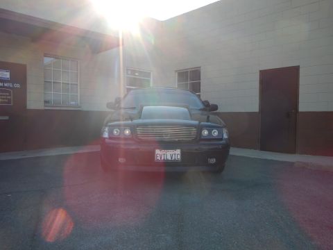 2003 Ford Crown Victoria - black full-size sedan parked head-on near a building sign reading Custom Mfg Co, backlit by the sun.
