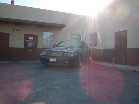 2003 Ford Crown Victoria - black full-size sedan parked on pavement beside a tan industrial building, backlit by strong sun and lens flare.
